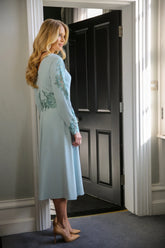 A woman wearing a long sleeve, midi length duck egg blue dress with embroidery detail at the waistline and sleeves.