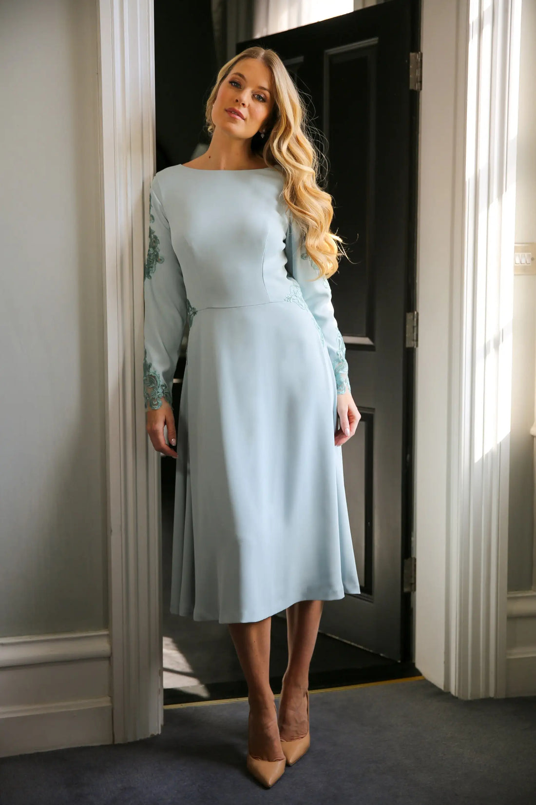 A woman wearing a long sleeve, midi length duck egg blue dress with embroidery detail at the waistline and sleeves.