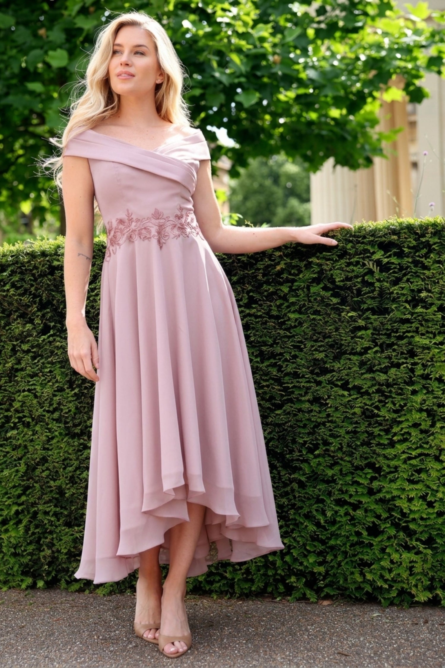 Woman in a pink off-shoulder dress standing against a green hedge.