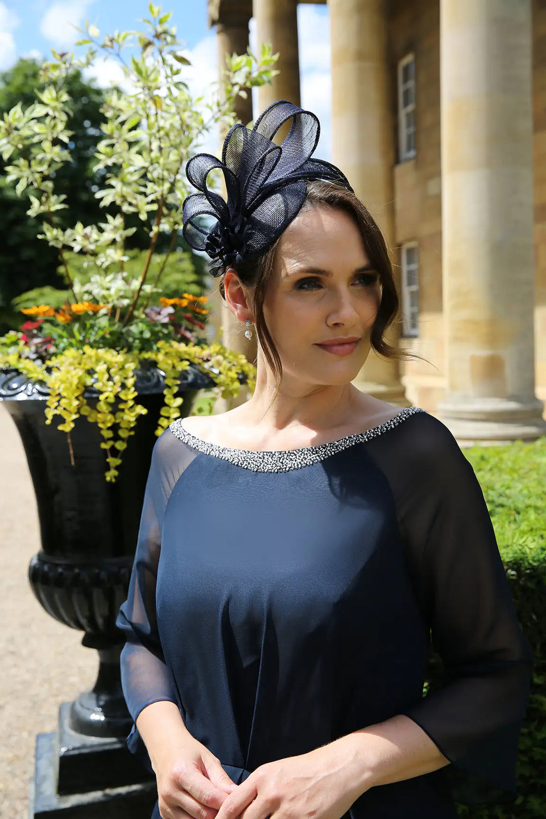 A woman wearing a navy fascinator with loops of sinamay and a small hand made flower to the right, positioned in front of a classical building.